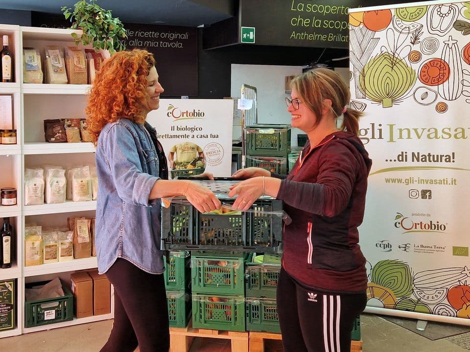 Due donne sorridono mentre si scambiano una cassetta di prodotti biologici all’interno di un negozio. Sullo sfondo scaffali con alimenti confezionati, cassette di verdura e un pannello promozionale dedicato agli ortaggi biologici.