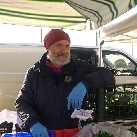 Ritratto di Flavio, mentre lavora al mercato protetto da una tenda parasole a strisce. Indossa un berretto rosso e guanti protettivi blu mentre sistema con cura verdura a foglia verde. L'espressione è serena e professionale.