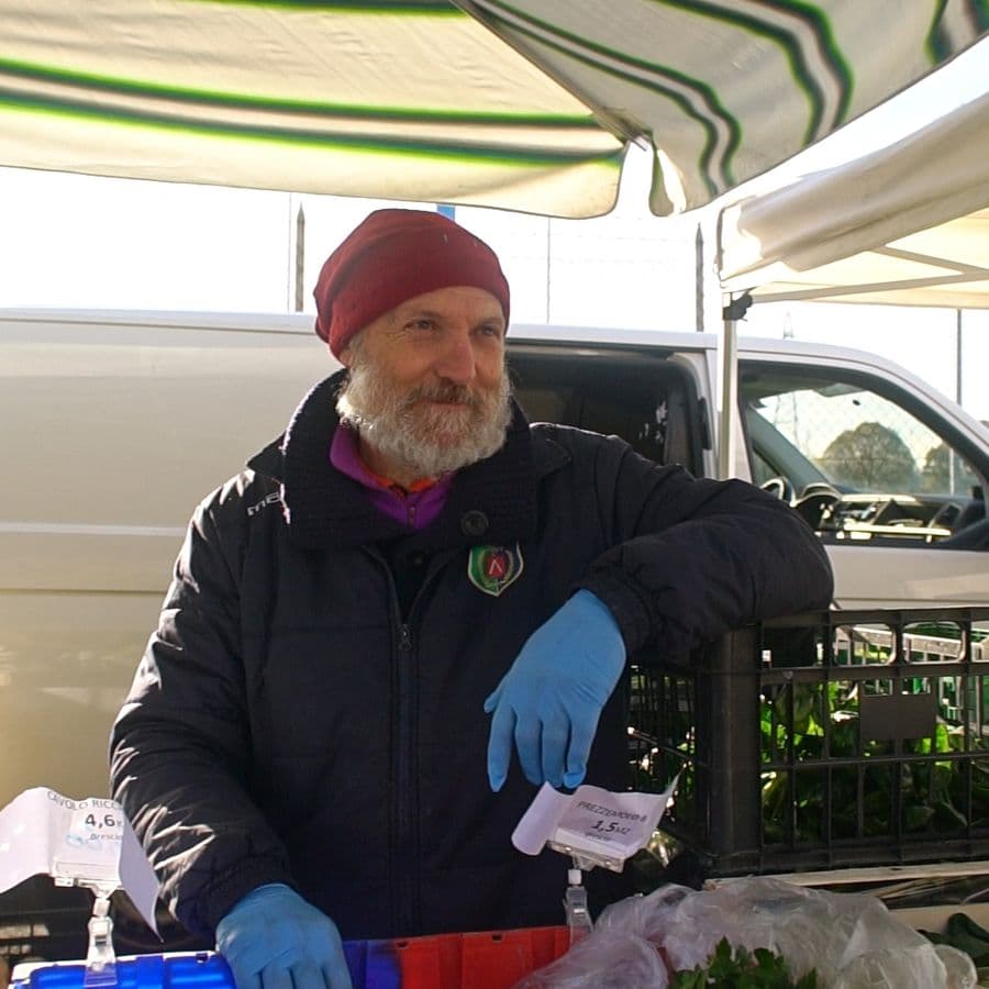 Ritratto di Flavio, mentre lavora al mercato protetto da una tenda parasole a strisce. Indossa un berretto rosso e guanti protettivi blu mentre sistema con cura verdura a foglia verde. L'espressione è serena e professionale.