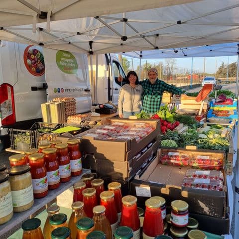 Inquadratura ravvicinata del banco del mercato "Cascina Maggia" che mette in risalto la varietà dei prodotti: in primo piano numerose bottiglie di passata di pomodoro, succhi di frutta e vasetti di conserve artigianali. Oltre le conserve si vedono cassette di fragole confezionate, ravanelli e uova fresche, con i due titolari che salutano cordialmente sullo sfondo.